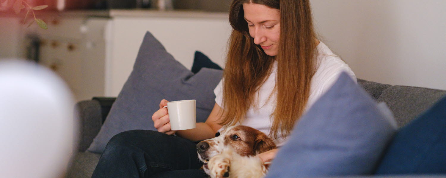 kvinna i soffa med hund i knät och kaffekopp i hand
