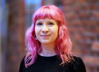 Cajsa i rosa hår och svart tröja framför en bakgrund av röd tegelvägg