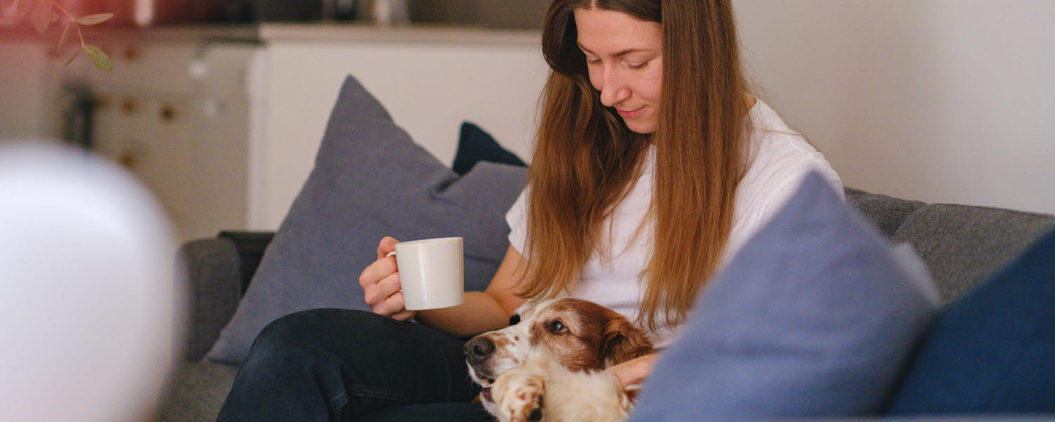 kvinna i soffa med hund i knät och kaffekopp i hand