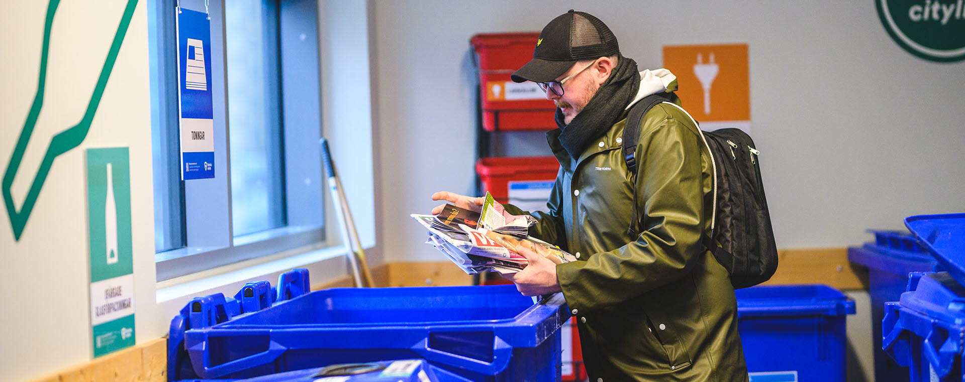 En man står i ett återvinningsrum och håller i en bunt tidningar ovanför ett blått avfallskärl.