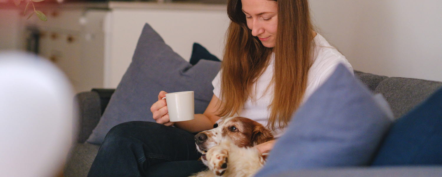 kvinna i soffa med hund i knät och kaffekopp i hand