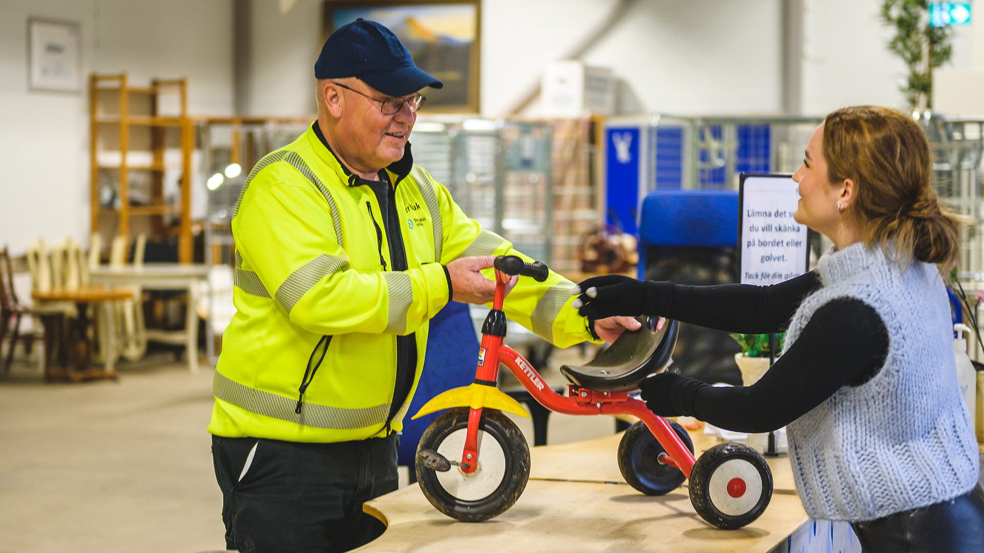 En kvinna lämnar in en liten trehjuling till återbruk, till en medarbetare på Återbrukshallen.