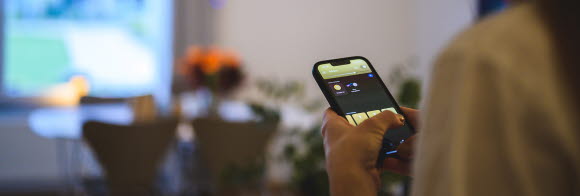 En person håller i en telefon där man ser en app för smart belysning. I bakgrunden syns en tänd lampa.