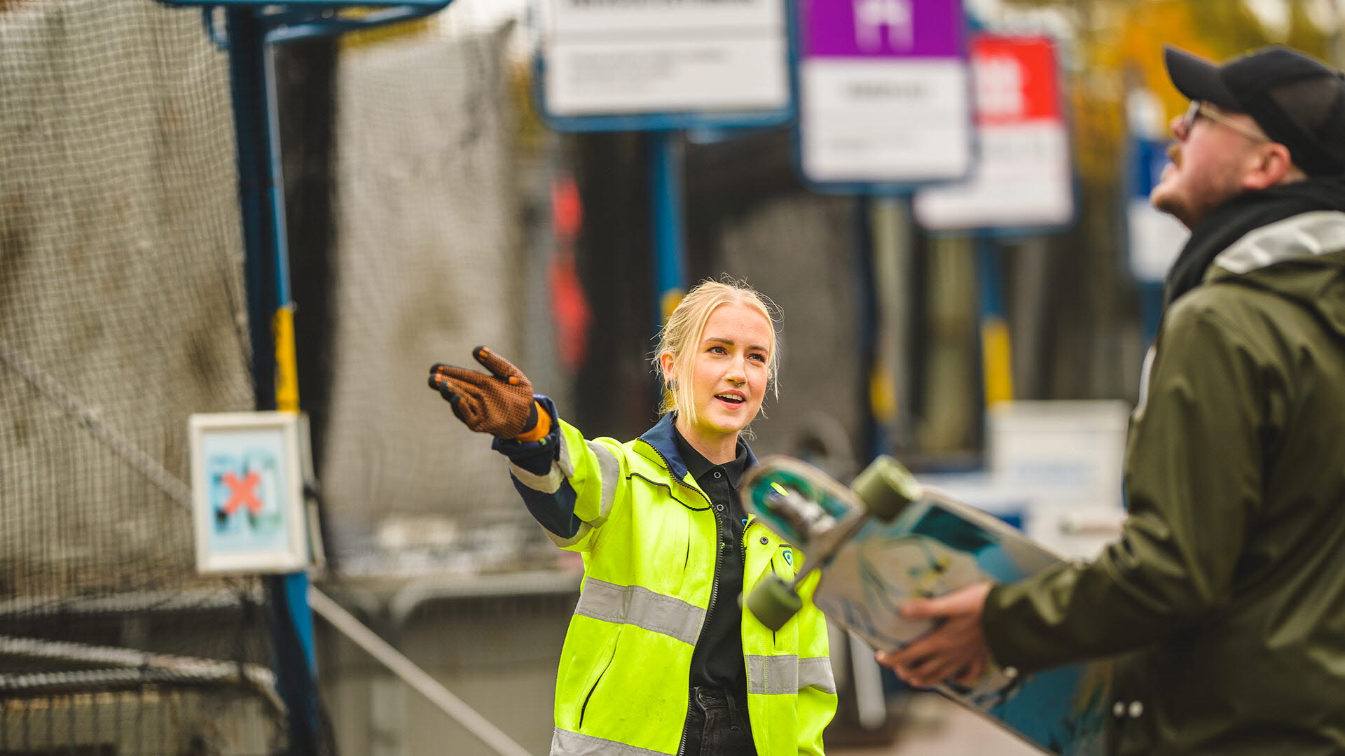 En medarbetare från Tekniska verken pekar mot en container. Bredvid henne står en kund med en skateboard i handen. 