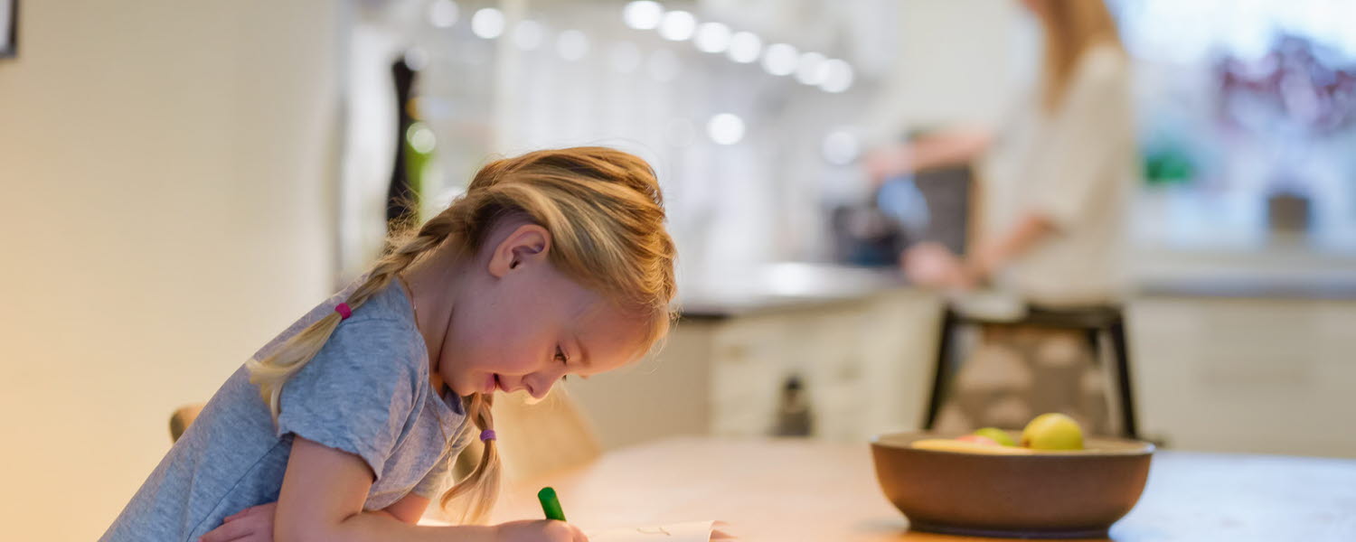 Flickar ritar vid ett bord medan mamma gör kaffe i bakgrunden